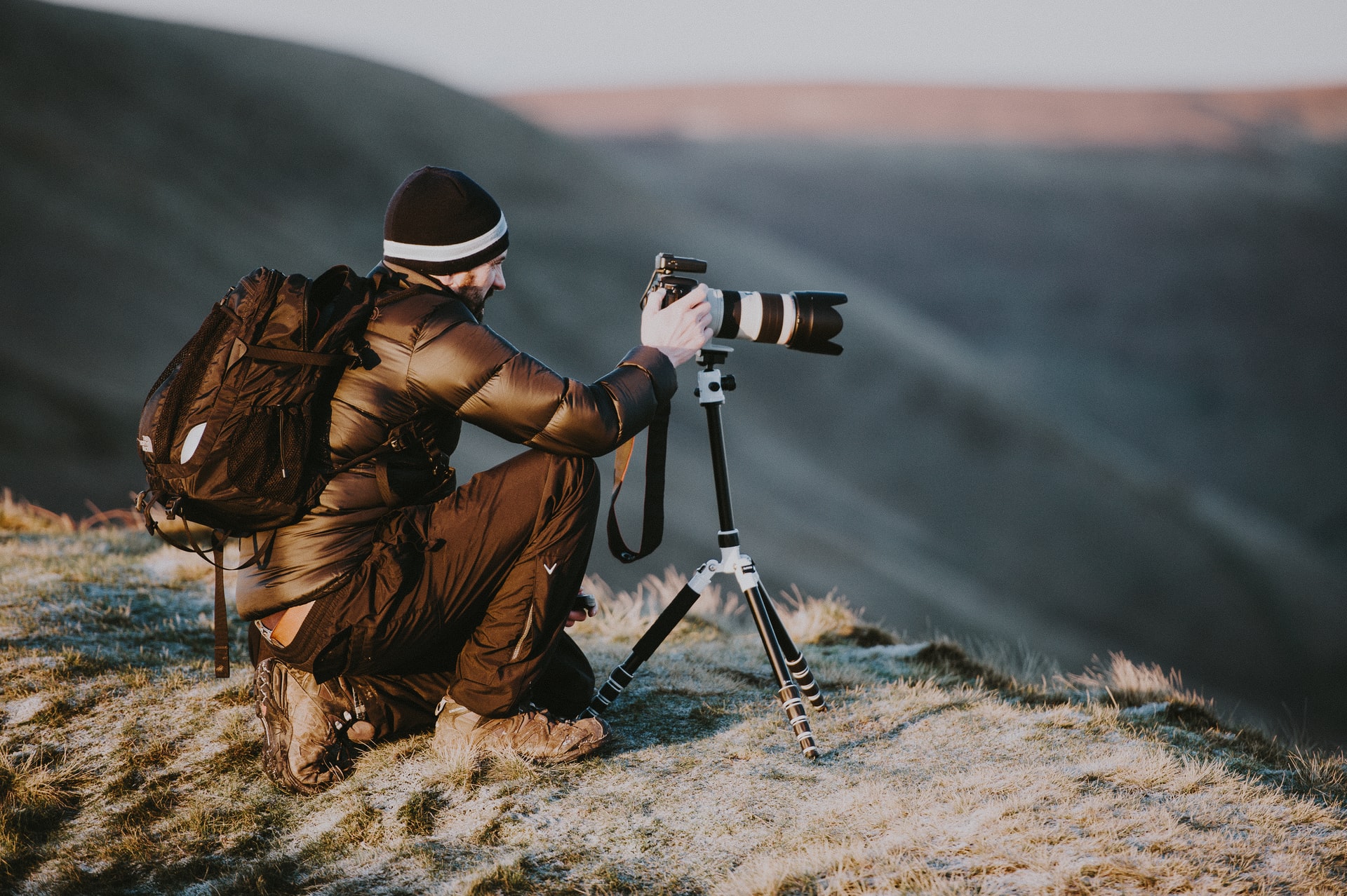  A stock photographer kneels on a grassy hilltop using a tripod to take a photo of a mountain range with the sun rising in the background.